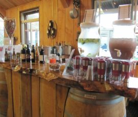 A rustic wooden bar holds wine bottles, drink dispensers, and cups, set atop two barrels inside a wooden room.