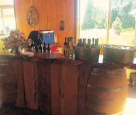 A rustic bar with wine bottles, coolers, and a floral arrangement set on wooden barrels inside a sunlit room.
