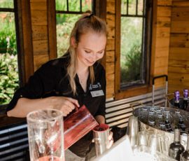 A woman pours a red drink from a pitcher into a metal cup at a bar with bottles and ice nearby.