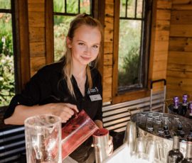 A woman behind a bar pours a drink from a pitcher into a cup, with bottles and glasses on the counter.