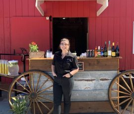 A woman stands in front of an outdoor bar setup with drinks, against a red barn backdrop on a sunny day.