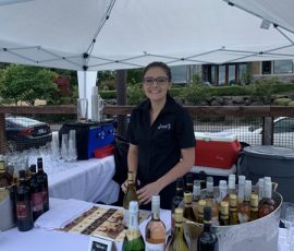 A woman stands behind a table with wine bottles and glasses under a white tent at an outdoor event.