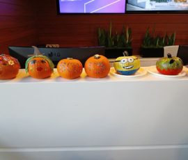 Six decorated pumpkins with various designs are displayed on a white counter in front of computer monitors and plants.