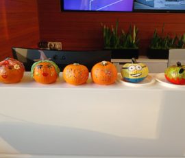 Five decorated pumpkins are displayed on a white counter in front of a desk and potted plants.