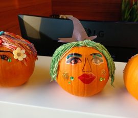 A pumpkin decorated with yarn hair, painted facial features, sequins, and a paper hat sits on a white surface.