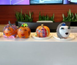 Five decorated pumpkins with painted faces and designs are displayed on a white counter in front of potted plants.