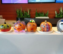 Six decorated pumpkins with various faces and designs are displayed on a white counter in front of green plants.
