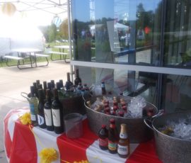 Bottles of wine, beer, and soda are arranged on a table with ice buckets at an outdoor event near a glass building.