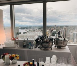 A table with wine bottles, glasses, and flowers is set up by large windows overlooking a city skyline and waterfront.