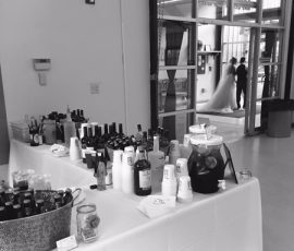 A table with assorted drinks and cups in the foreground; a bride and groom stand together in the background.
