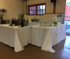 A bar setup with bottles, ice buckets, glasses, and a “BAR” sign on white tablecloth-covered tables indoors.