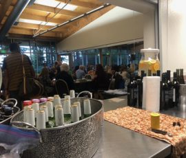 Beverages on a counter in the foreground with a group of people seated at tables in a meeting room in the background.