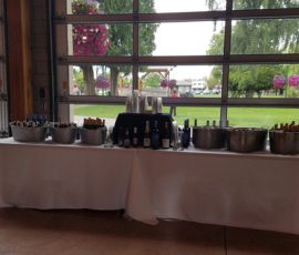 Table with buckets of bottled drinks and wine bottles set up indoors near large windows overlooking a green outdoor area.