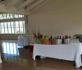 A table with drinks, glasses, and flowers set up in a spacious room with large windows and another table in the background.