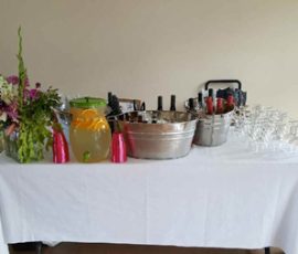 Table with a white cloth holding a floral arrangement, glass drink dispenser, ice buckets with bottles, and stacked glasses.
