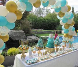 A dessert table with blue and gold treats stands under a balloon arch by a pool at an outdoor party.