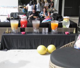 A table with four beverage dispensers, cups, and cones is set up near seating at an outdoor gathering.