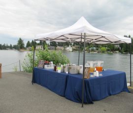 A white canopy tent with tables covered in blue cloth holds drinks and supplies beside a lake under a cloudy sky.