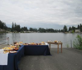 Tables with food and drinks set up by a lakeside on a cloudy day, with houses and trees visible across the water.