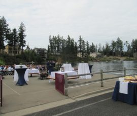 Outdoor event setup by a river with tables covered in white and navy cloths; people seated at left.