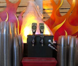 Two beer taps between stacks of clear plastic cups, with a fiery flame backdrop and wooden panels behind.