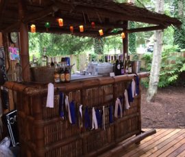 Outdoor tiki bar with various bottles, string lights, and blue and white ribbons, surrounded by trees and wooden deck.