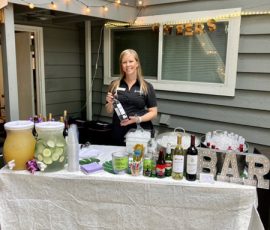 A woman stands behind an outdoor bar table set with drinks, mixers, and wine bottles, with "CHEERS" and "BAR" signs.