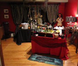 A home bar decorated for Halloween with bottles, a skeleton, a doll, and spooky props in a dimly lit room.