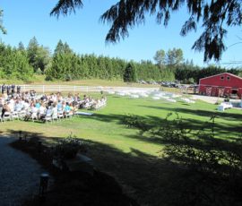 Outdoor event with people seated at tables on a lawn near a red barn, surrounded by trees and a white fence.