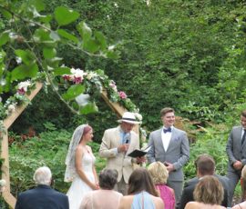 A bride and groom stand with an officiant and groomsman during an outdoor wedding ceremony with guests seated.