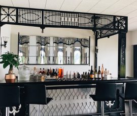 Modern home bar with black stools, assorted bottles, a plant, and a large mirror behind the counter.