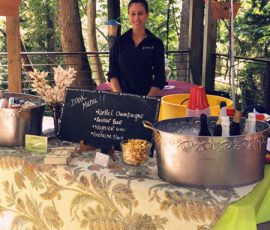 Woman stands behind an outdoor drink station with a menu board, ice buckets, snacks, and drink supplies on the table.