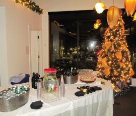 A decorated table with drinks sits near a lit Christmas tree in a room with festive lighting and large windows.
