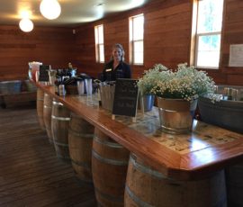 A woman stands behind a bar made of wooden barrels, with drinks and flowers set up in a rustic wooden room.