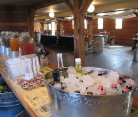 Beverage station with iced bottled drinks, pitchers of juice, and plastic cups set up in a rustic wooden event hall.
