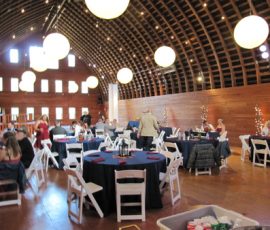 People sit at round tables with black tablecloths in a large, wooden event hall with hanging lights and high arched ceiling.
