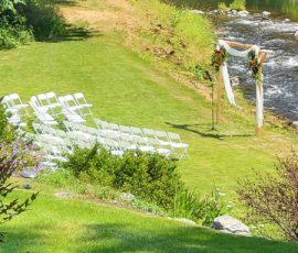 White folding chairs are arranged on grass facing a decorated arch near a river, set up for an outdoor ceremony.