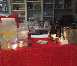 A table with drinks, wine bottles, a lemon water dispenser, and festive decorations, set in front of shelves.