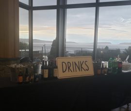A table with assorted beverages and a sign labeled "DRINKS" set by large windows overlooking a scenic outdoor view.