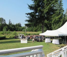 Outdoor wedding ceremony with guests seated on lawn near a white tent, surrounded by trees and greenery.