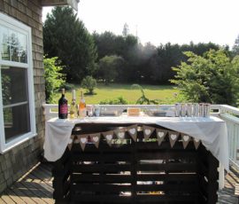 Outdoor bar setup on a wooden deck with bottles, cans, and drinks, overlooking a grassy yard and trees.