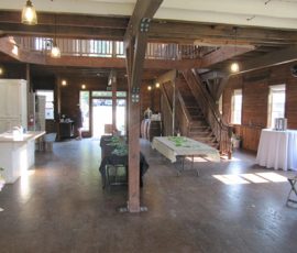 Spacious wooden interior with exposed beams, tables set up, and a staircase leading to an upper loft area.