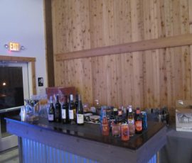 A wooden bar counter with various bottles of wine, spirits, and mixers, set against a wooden wall inside a venue.