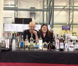 Two people stand behind a table displaying various bottles of alcohol in an industrial setting.