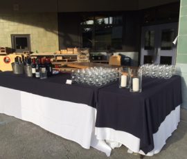 A table covered with a black and white cloth holds wine bottles, glasses, candles, and ice buckets outside a building.