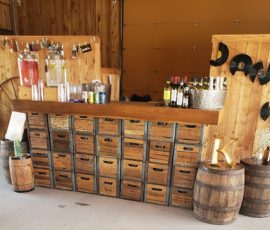 Rustic bar setup with wooden crates, barrels, assorted bottles, and hanging keys against wooden walls.