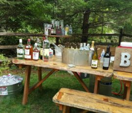 Outdoor bar setup with various liquor bottles, mixers, ice buckets, and a metal "BAR" sign on wooden tables.