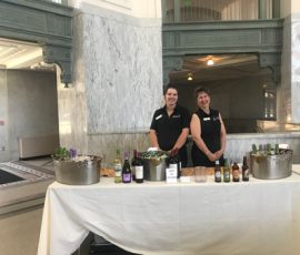 Two women stand behind a table with wine bottles, beer, and ice buckets in a large, ornate indoor space.