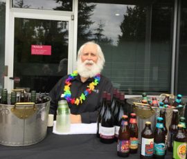 An older man with a white beard sits behind a table with bottles of wine and beer, wearing a colorful lei.