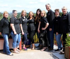 Eight women and one man, all in matching black shirts, stand side by side on an outdoor patio with plants in the background.